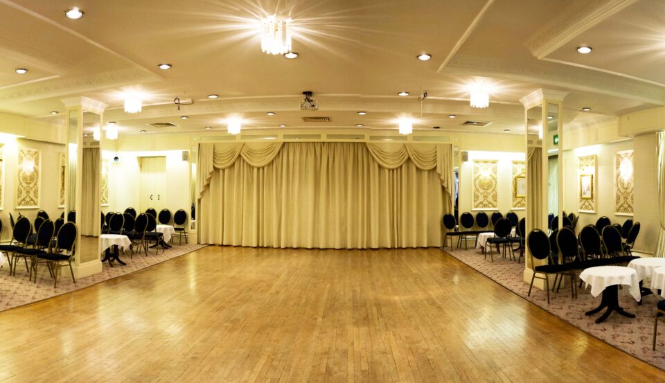 A Ballroom with wooden floor down the centre and carpet along the right and left sides. Carpeted area has tables and chairs set up.