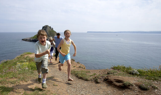 A young boy and girl running along the cliff with their parents following and Thatcher Rock and the sea in the distance.